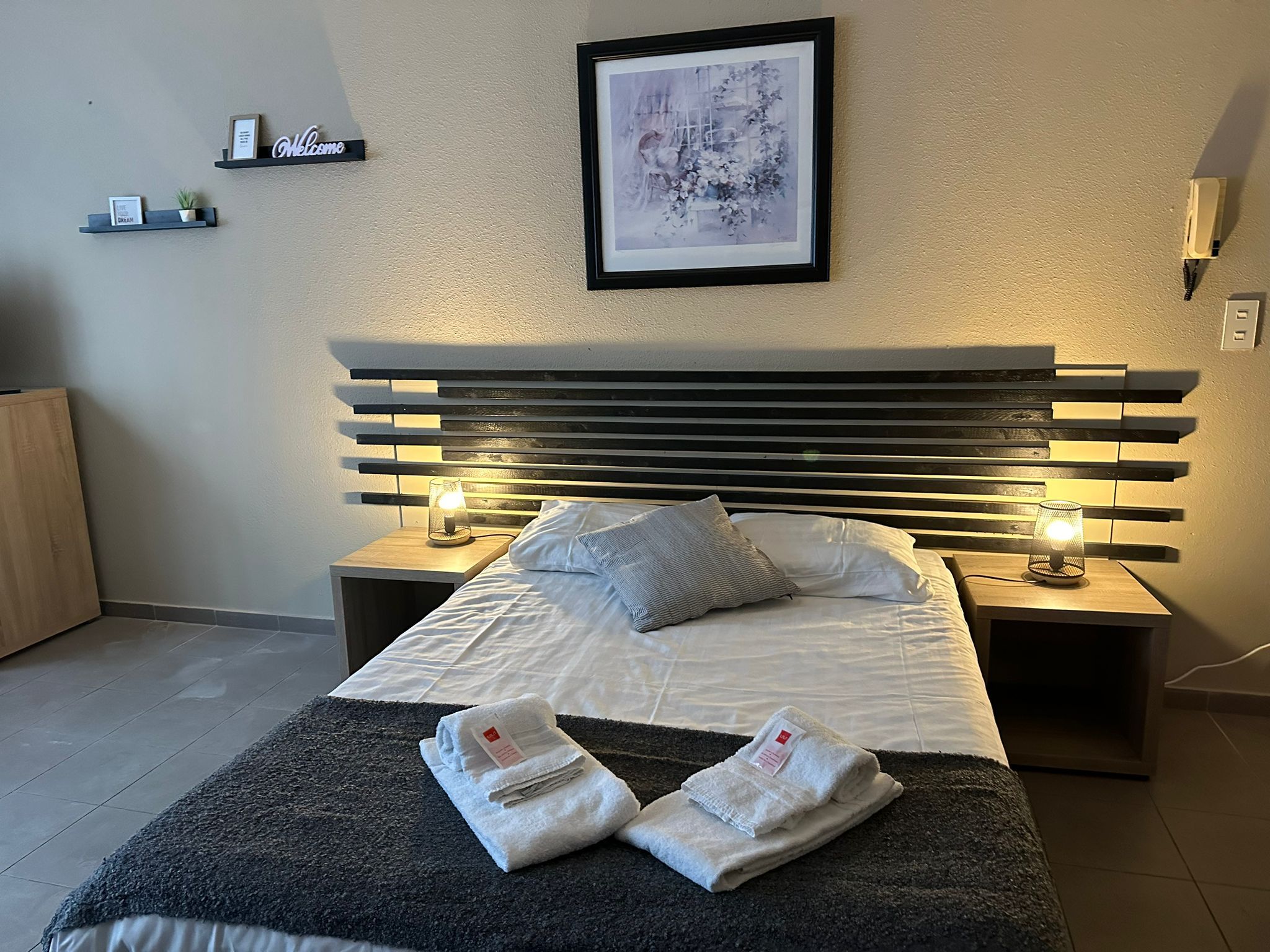 A neatly made bed with a headboard, two bedside tables with lamps, a framed picture on the wall, and a small shelf with a welcome sign.