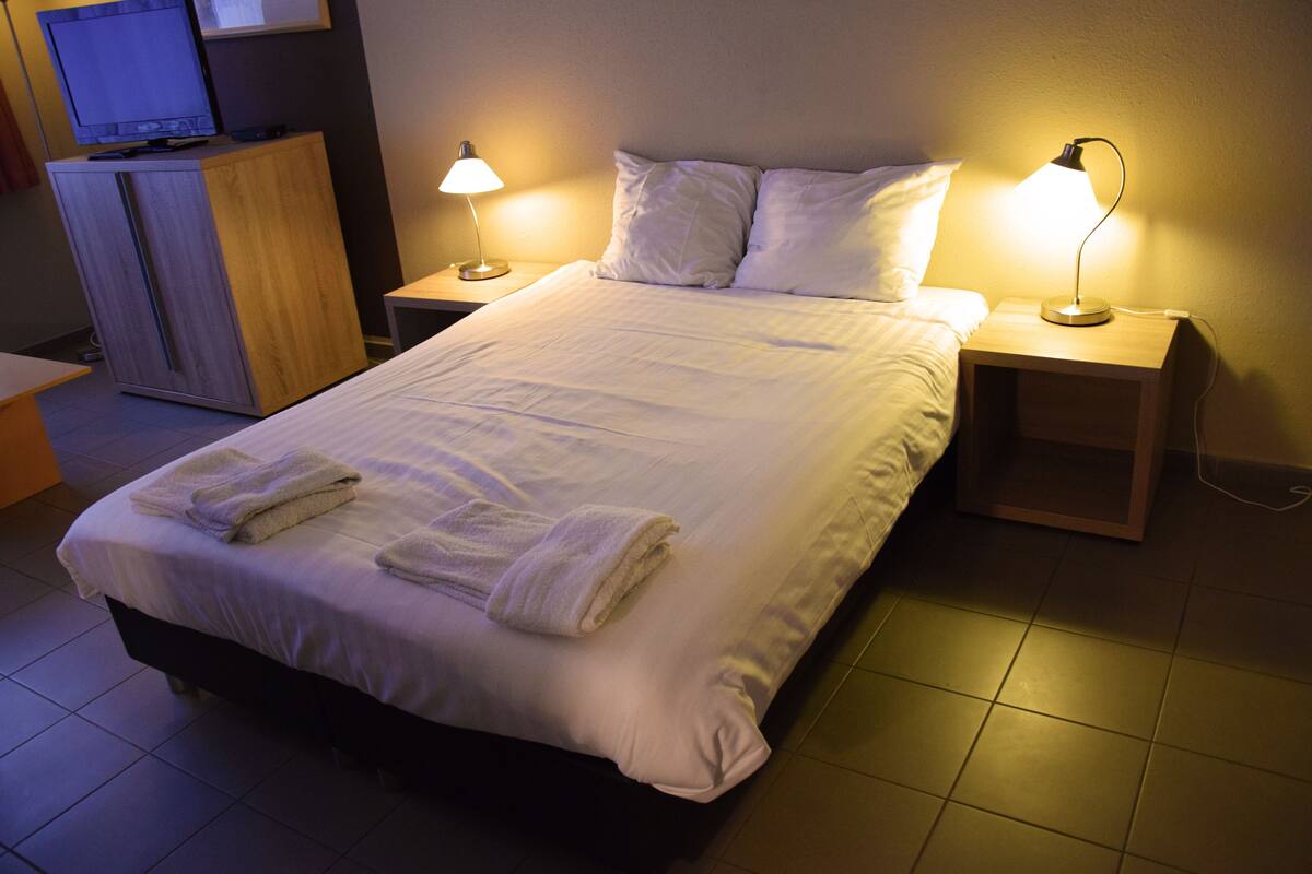 A neatly made bed with white linens and towels, two bedside lamps, a wooden nightstand, and a television on a cabinet.