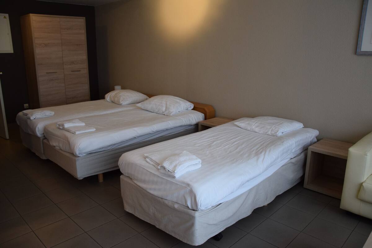 A hotel room with two single beds, each with a pillow and a folded towel. There is a wooden wardrobe and a small wooden nightstand with a lamp.