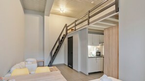 A compact hotel room with a bed, a small table, a sofa, a staircase, and a kitchenette. - Budget Flats Leuven (Leuven)