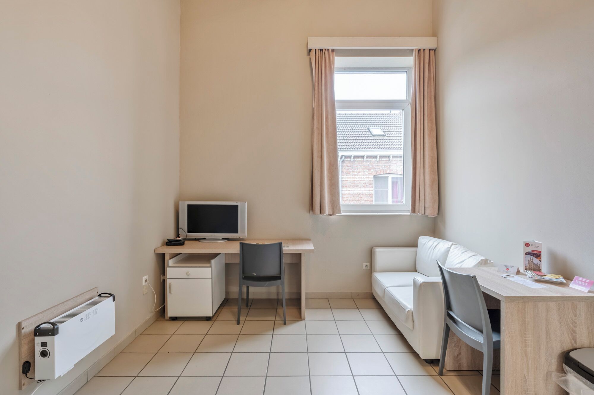 Une petite pièce bien éclairée, avec un bureau, une chaise, un canapé blanc et une fenêtre avec des rideaux.