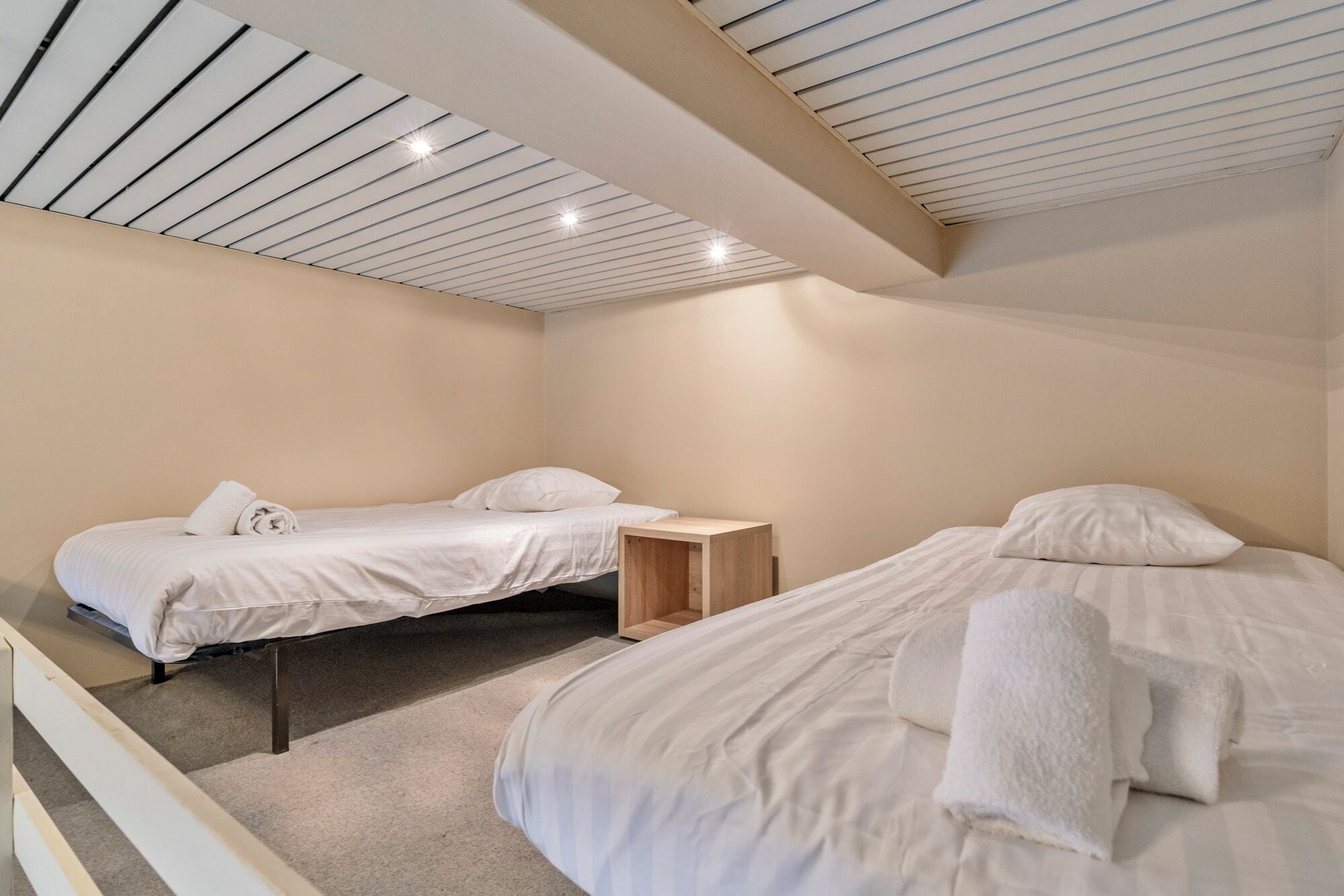 Une chambre d’hôtel avec deux lits, une petite table de chevet en bois et un plafond en pente doté d’un éclairage encastré.