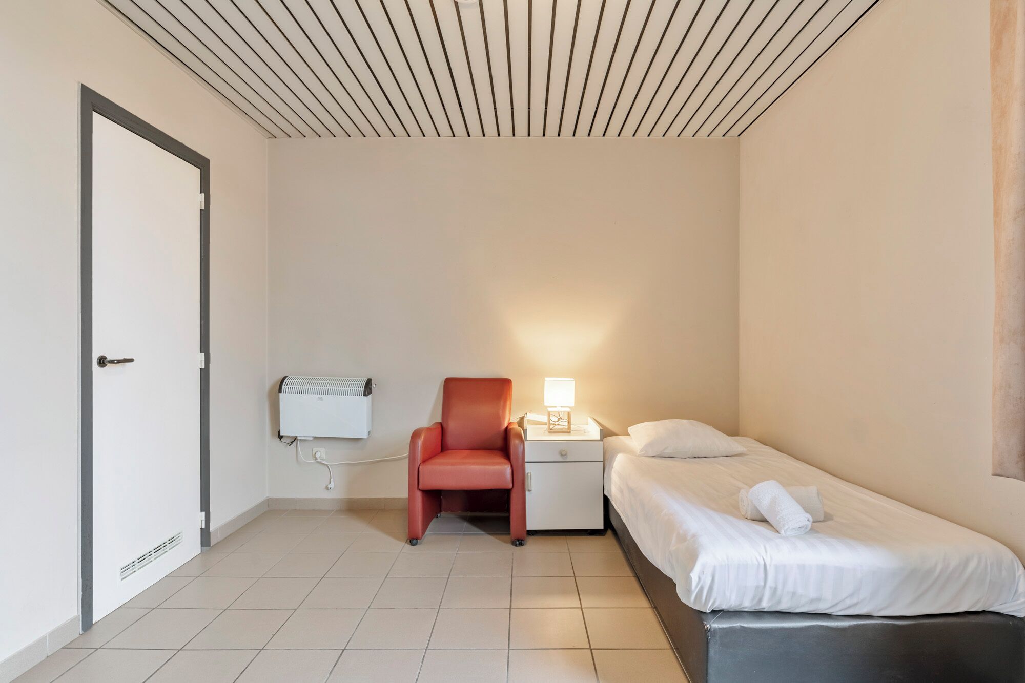 A small, clean room with a bed, a chair, a nightstand with a lamp, and a wall-mounted heater.