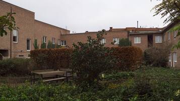Un immeuble résidentiel aux murs de briques, doté de nombreuses fenêtres et comprenant un espace jardin avec un banc et diverses plantes.