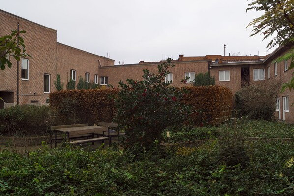 Un edificio residencial con paredes de ladrillo, múltiples ventanas y un jardín con un banco y diversas plantas.