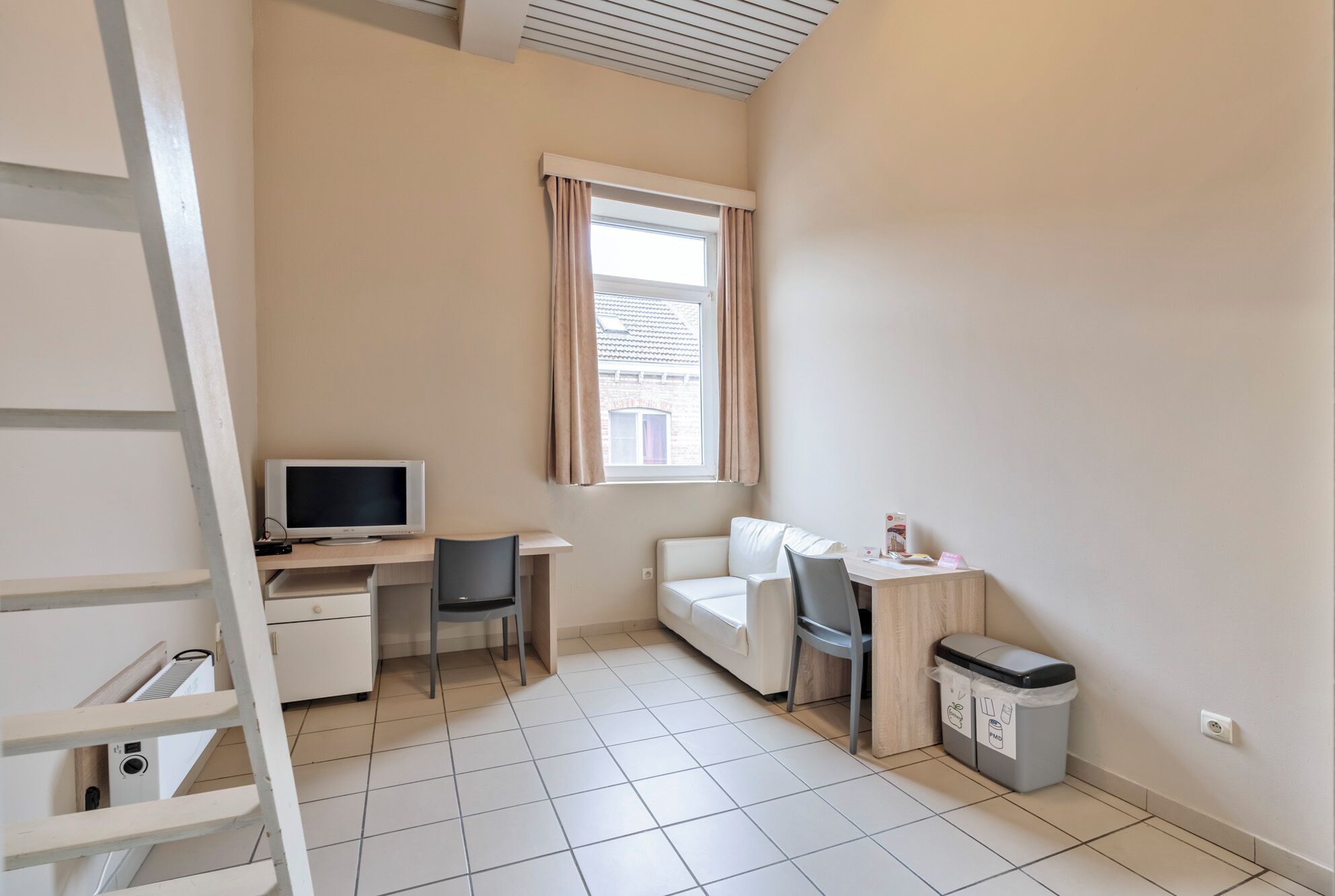 A small, well-lit room with a desk, a chair, a sofa, and a TV.