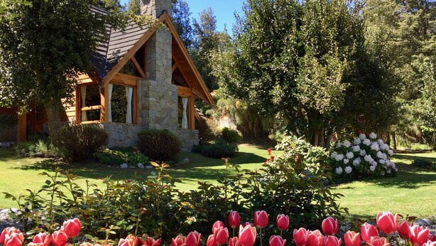 A stone cottage with a sloped roof, surrounded by a well-maintained garden with red tulips and white flowers. - Tillka, Casas de Montaña (Villa La Angostura)