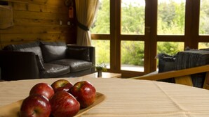 A wooden cabin interior with a leather sofa, a wooden table with apples, and large windows with a view of greenery. - Tillka, Casas de Montaña (Villa La Angostura)
