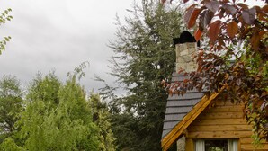 A rustic cabin with a stone chimney, a wooden deck, and a gravel path leading to a white chair. - Tillka, Casas de Montaña (Villa La Angostura)