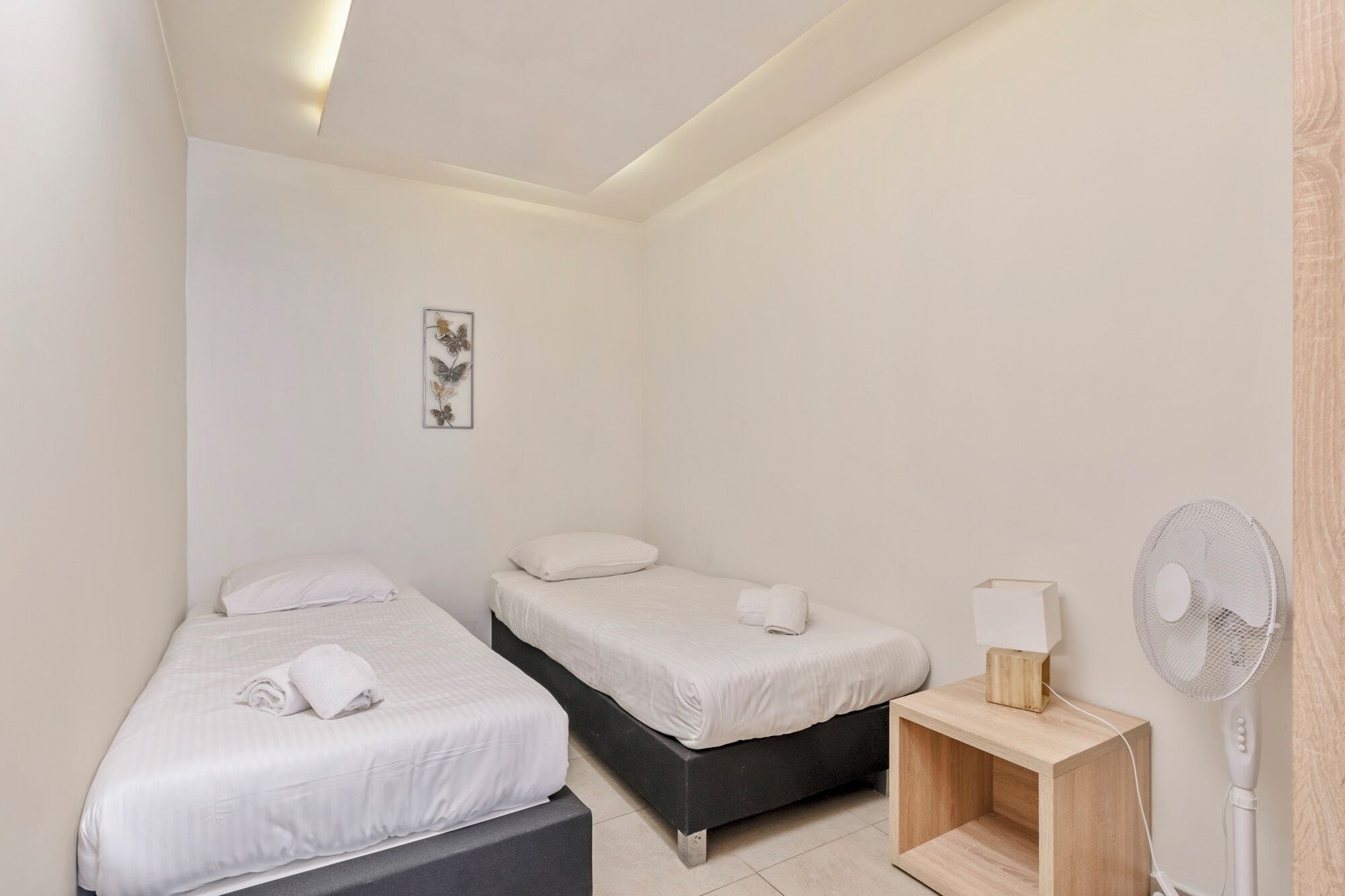Habitación de hotel con dos camas, una mesita de noche con lámpara y un ventilador de pie.