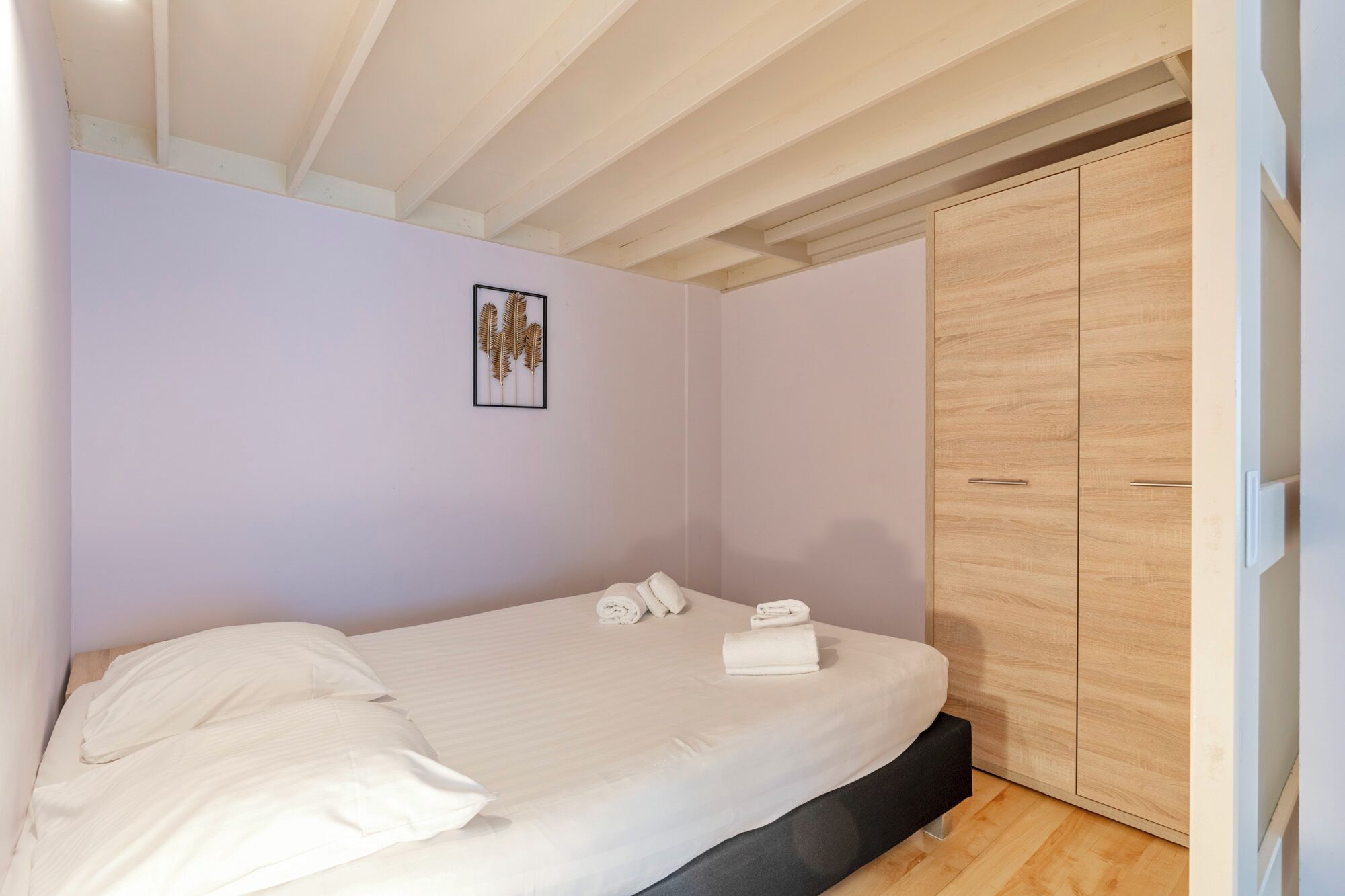 A bedroom with a bed, a wooden wardrobe, and a framed artwork on the wall.