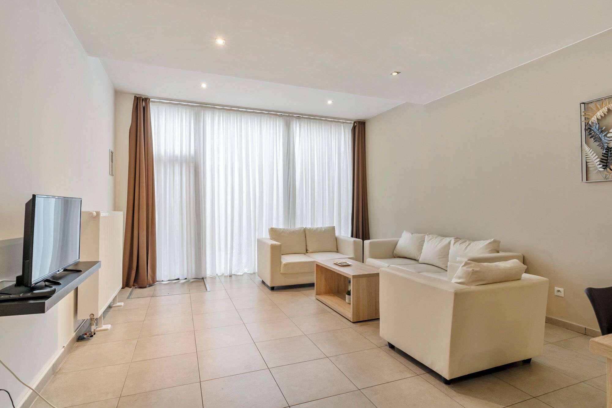 A modern living room with a flat-screen TV, two sofas, a coffee table, and a large window with curtains.