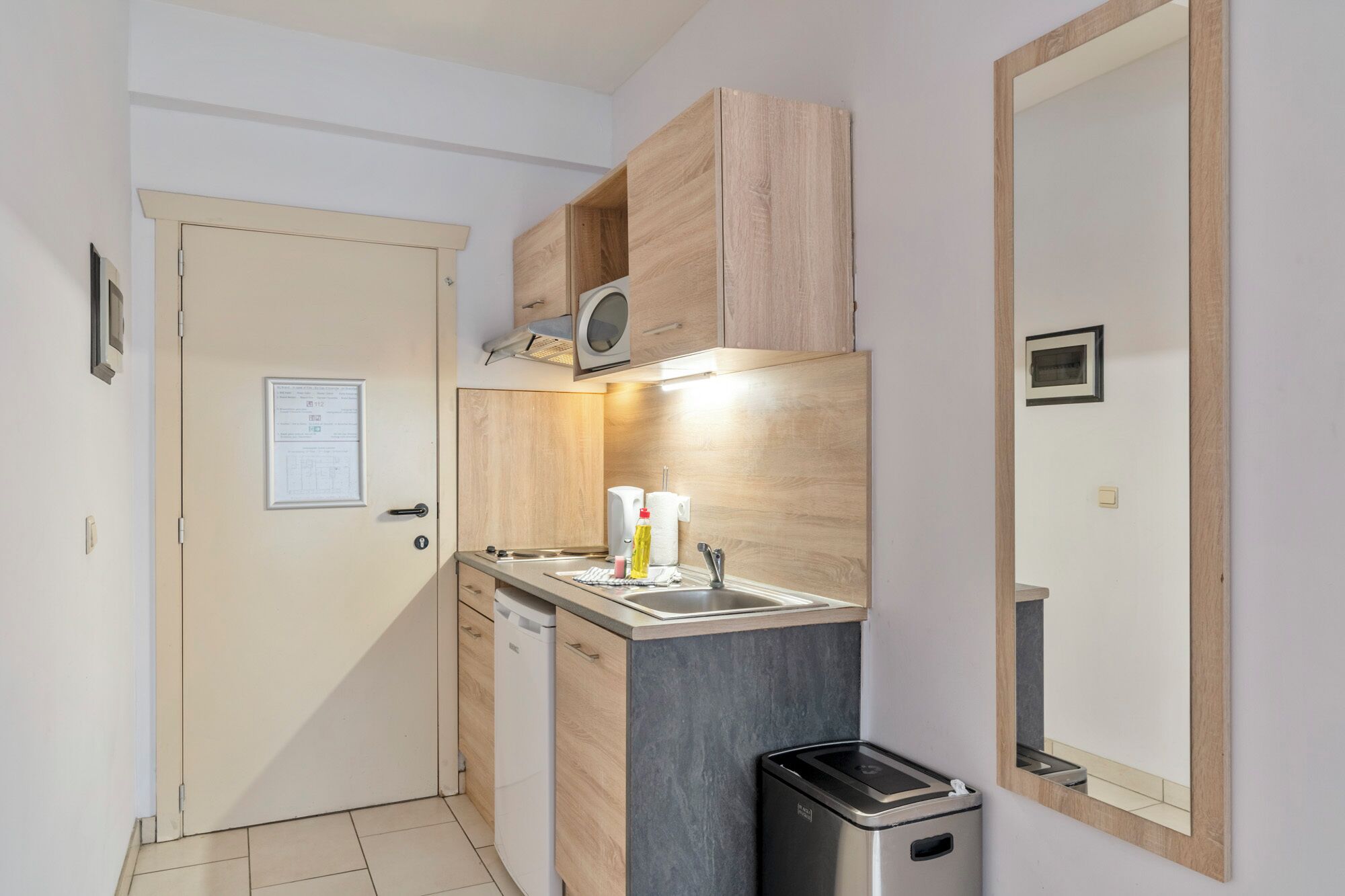 A compact kitchen with a microwave, sink, and a trash bin. A door with a notice is visible, and the room has a tiled floor.