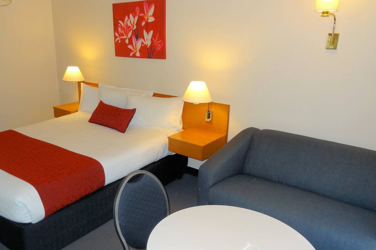 A hotel room with a bed, two bedside lamps, a red throw blanket, a grey sofa, and a small round table.
