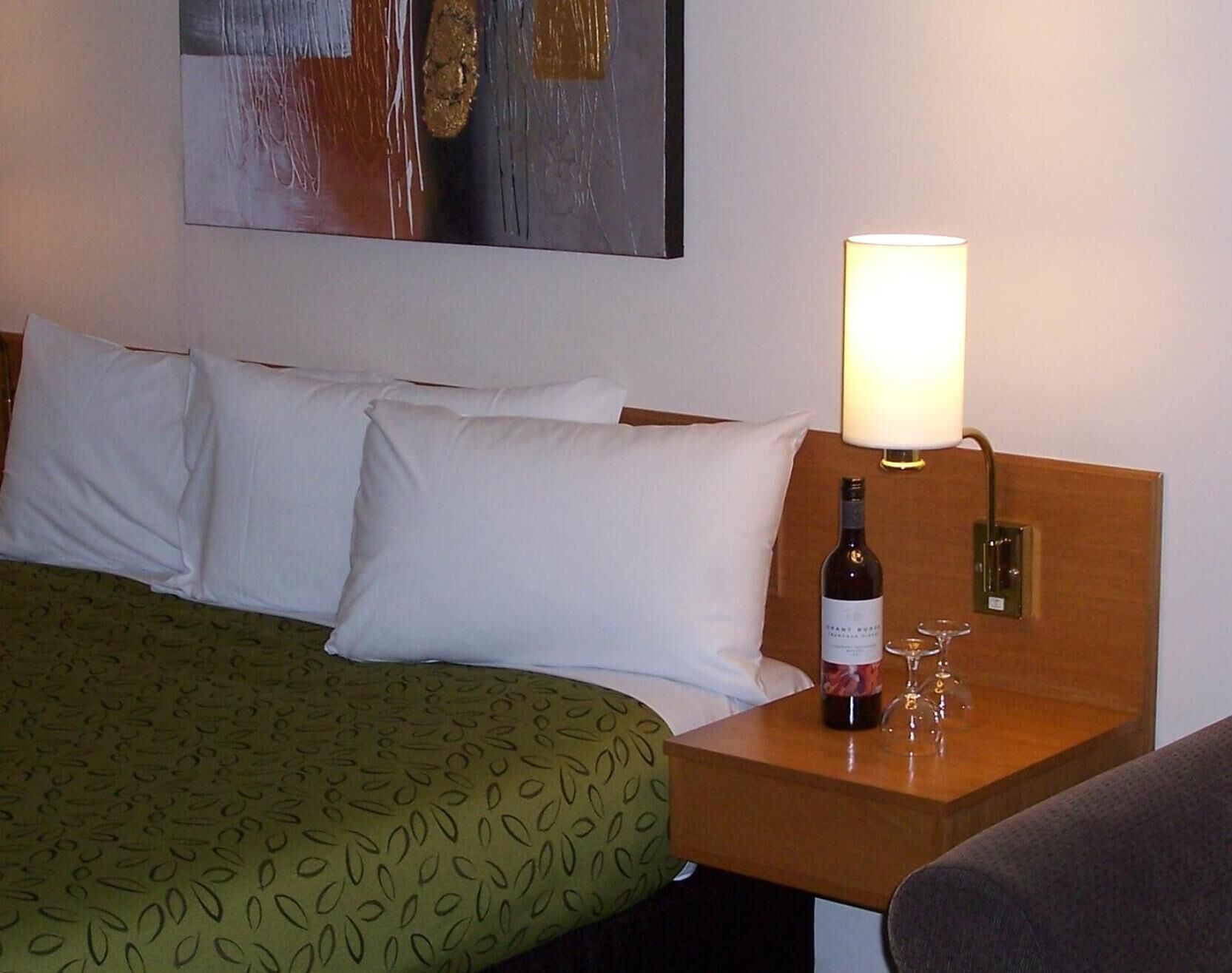 Camera d'albergo con un letto, cuscini, copripiumino verde fantasia, un comodino in legno con una bottiglia di vino e bicchieri, e una lampada a parete.