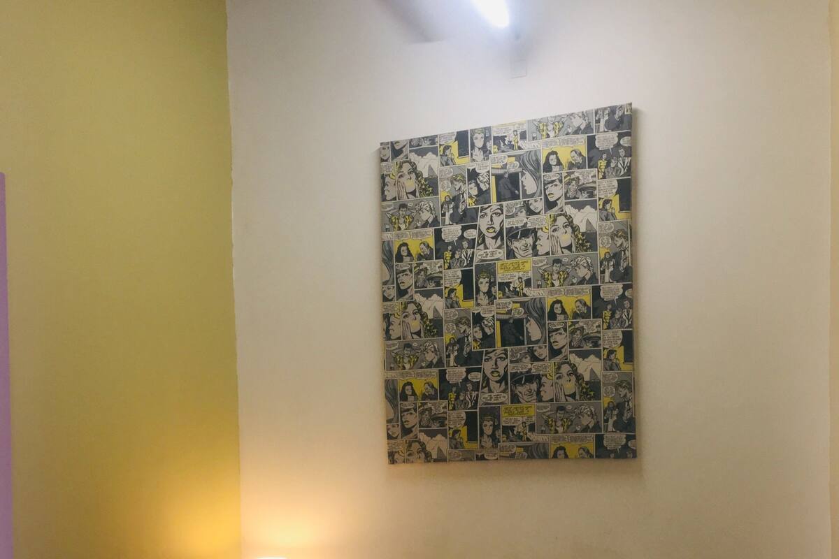 A hotel room with a bed, a wall-mounted comic strip artwork, a nightstand with a lamp, and a speaker.