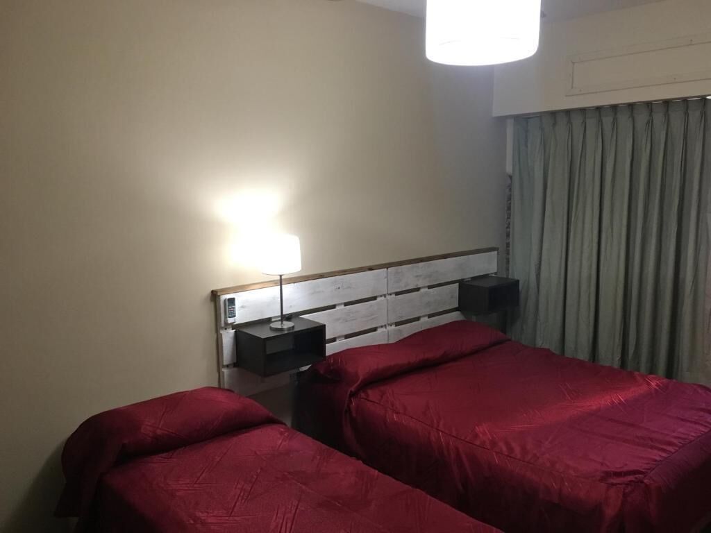 A hotel room with two beds, a bedside table with a lamp, and a window with curtains.