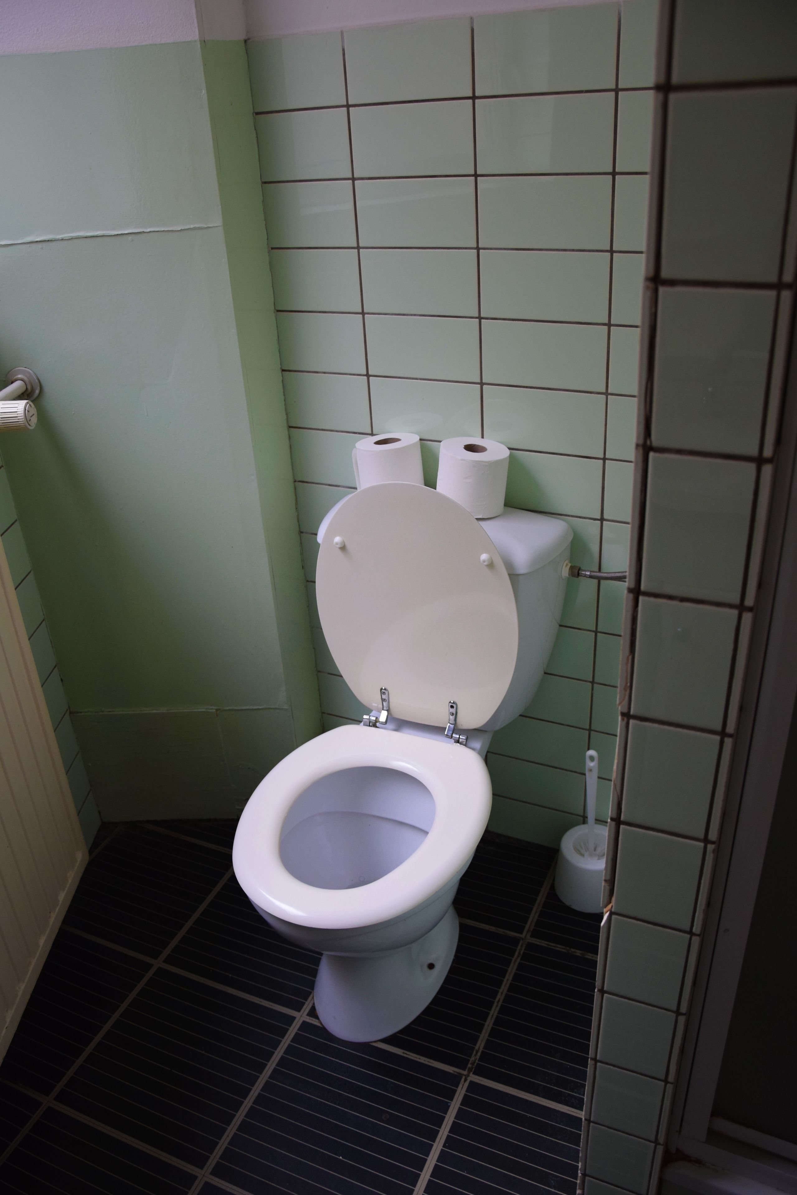 A bathroom with a toilet, two rolls of toilet paper, and a plunger on the floor.