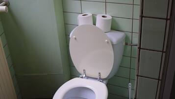 A bathroom with a toilet, two rolls of toilet paper, and a plunger on the floor.