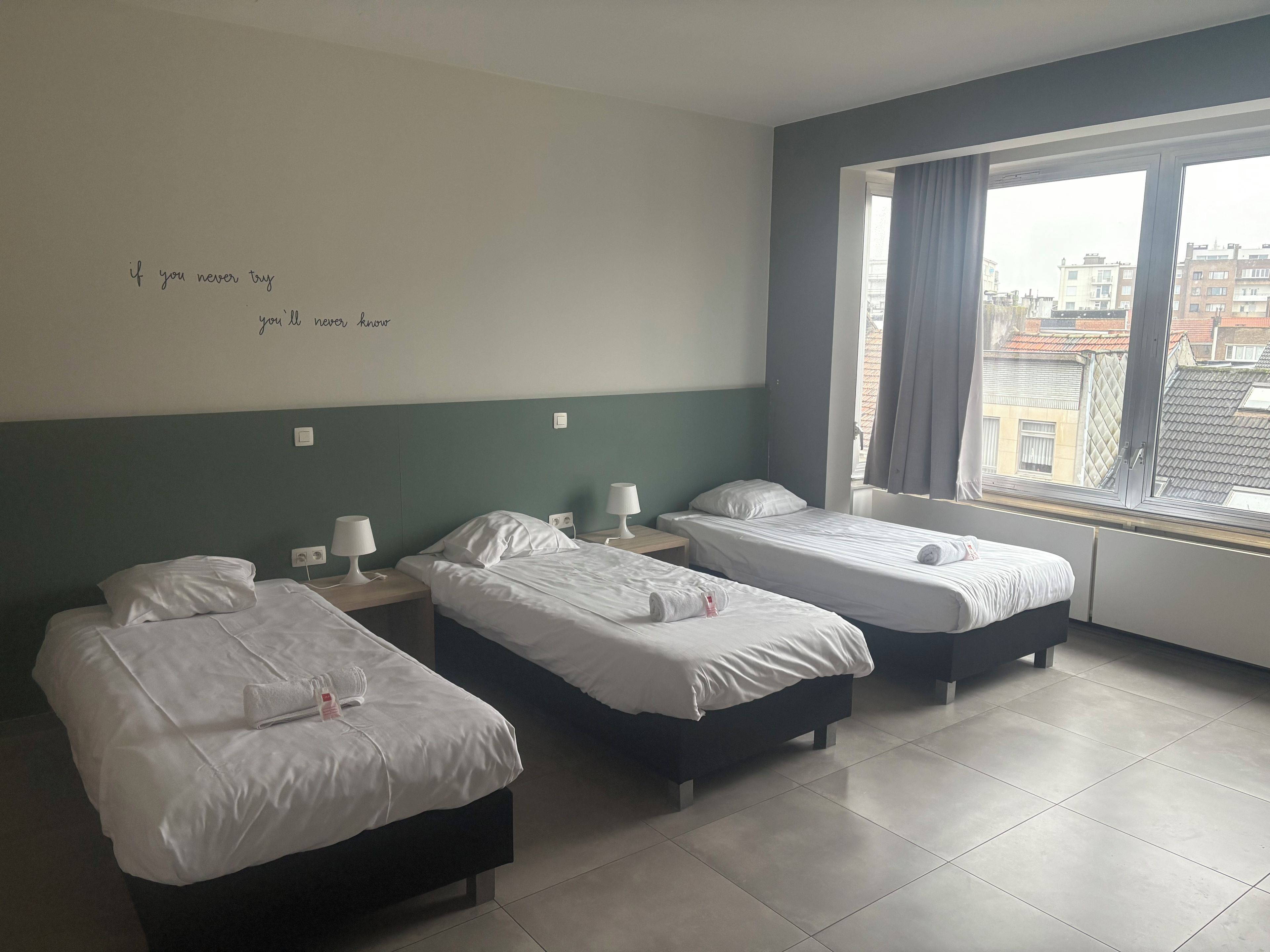 A hotel room with three beds, a window with curtains, and a wall with a motivational quote.