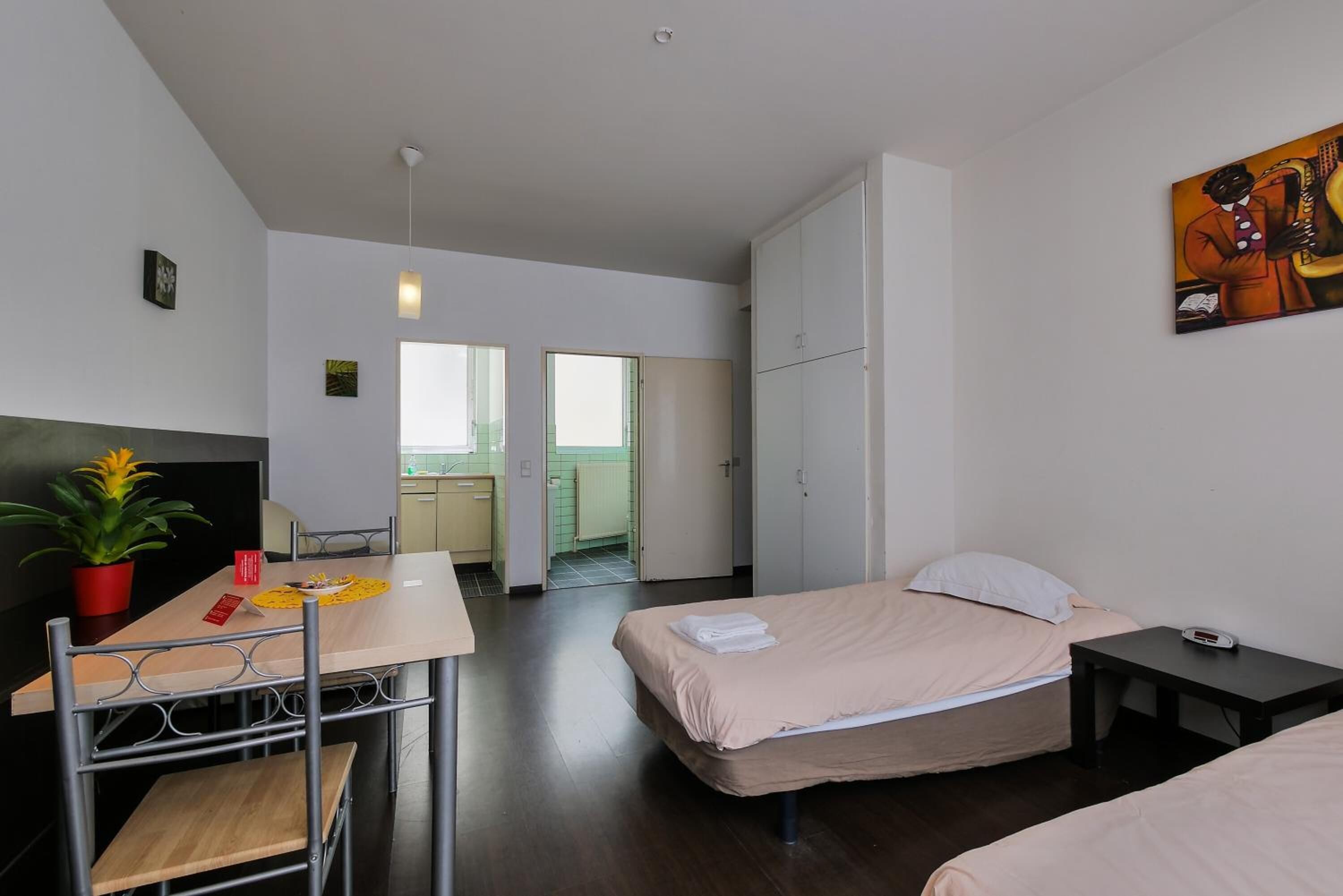 A modern hotel room with a bed, bedside table, and a desk with a chair. There is a potted plant on the desk, and a painting on the wall.