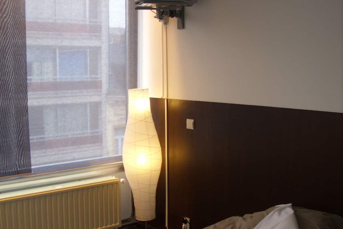 A hotel room with a bed, a TV mounted on the wall, a lamp, and a window with a view of buildings.