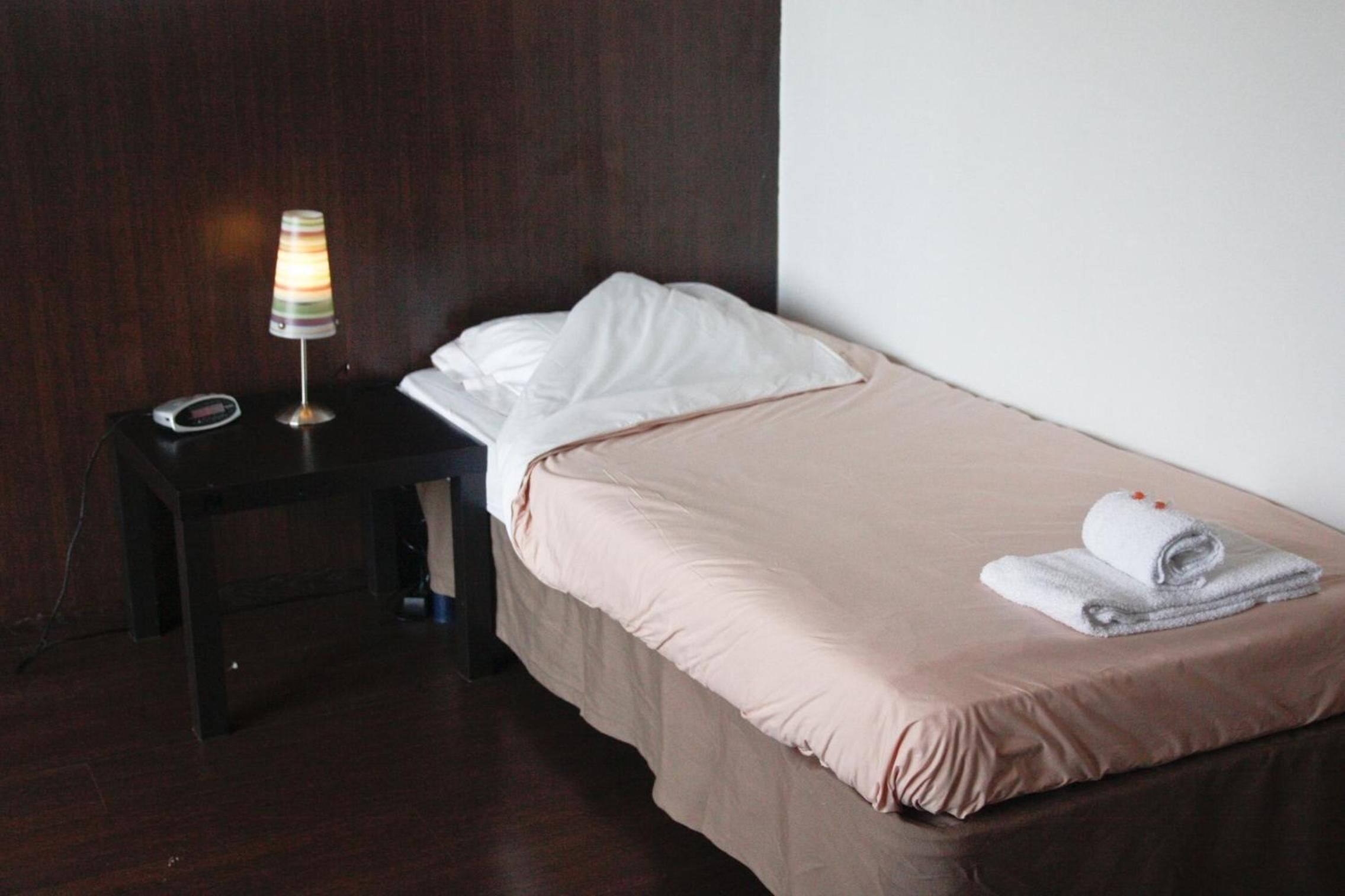Een enkel bed met een beige dekbedovertrek en witte kussens, een klein nachtkastje met een lamp, en een opgevouwen handdoek op het bed.