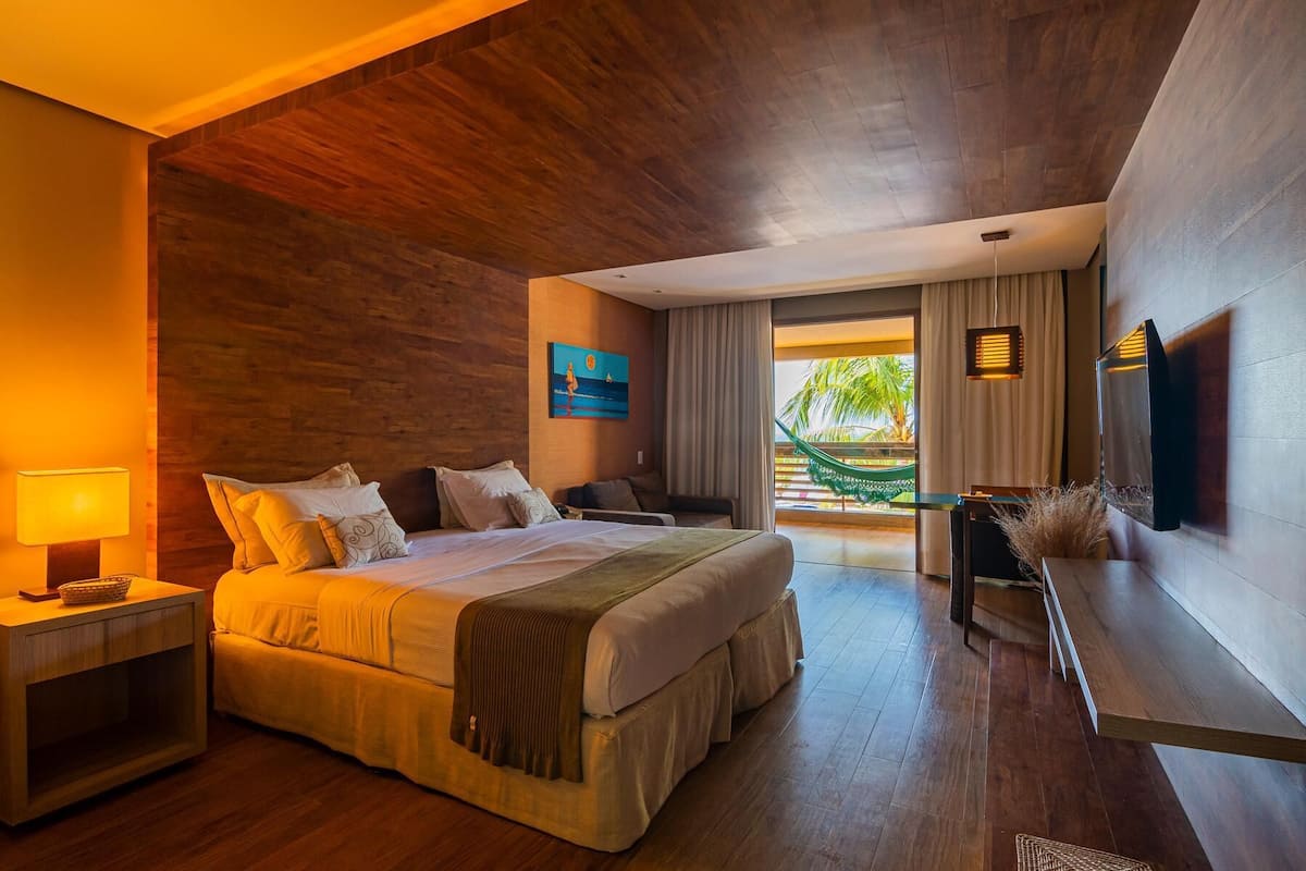 Habitación de hotel con cama, mesitas de noche, escritorio, televisor y vistas a un balcón con plantas.