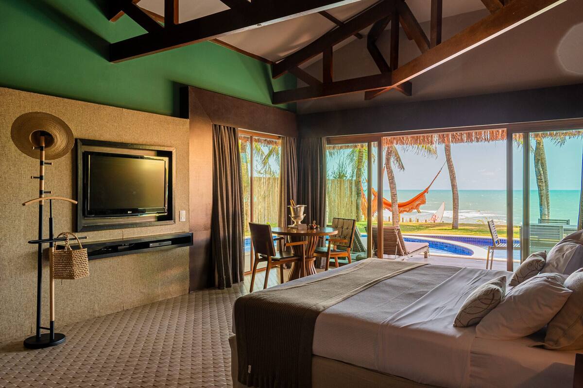 Habitación de hotel con una cama grande, televisión montada en la pared, zona de comedor con mesa y sillas, y vistas al océano a través de ventanas amplias.