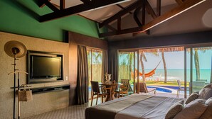Habitación de hotel con una cama grande, televisión montada en la pared, zona de comedor con mesa y sillas, y vistas al océano a través de ventanas amplias.