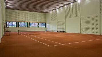 Una cancha de tenis interior con superficie de arcilla, rodeada por paredes con paneles perforados y un techo de madera con luminarias.