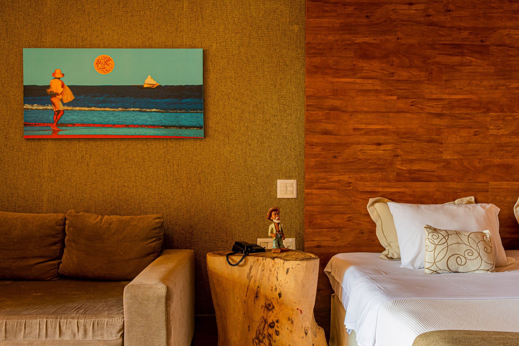 A room with a wooden wall, a painting of a beach scene, a wooden nightstand with a telephone, and a bed with white bedding and patterned pillows.