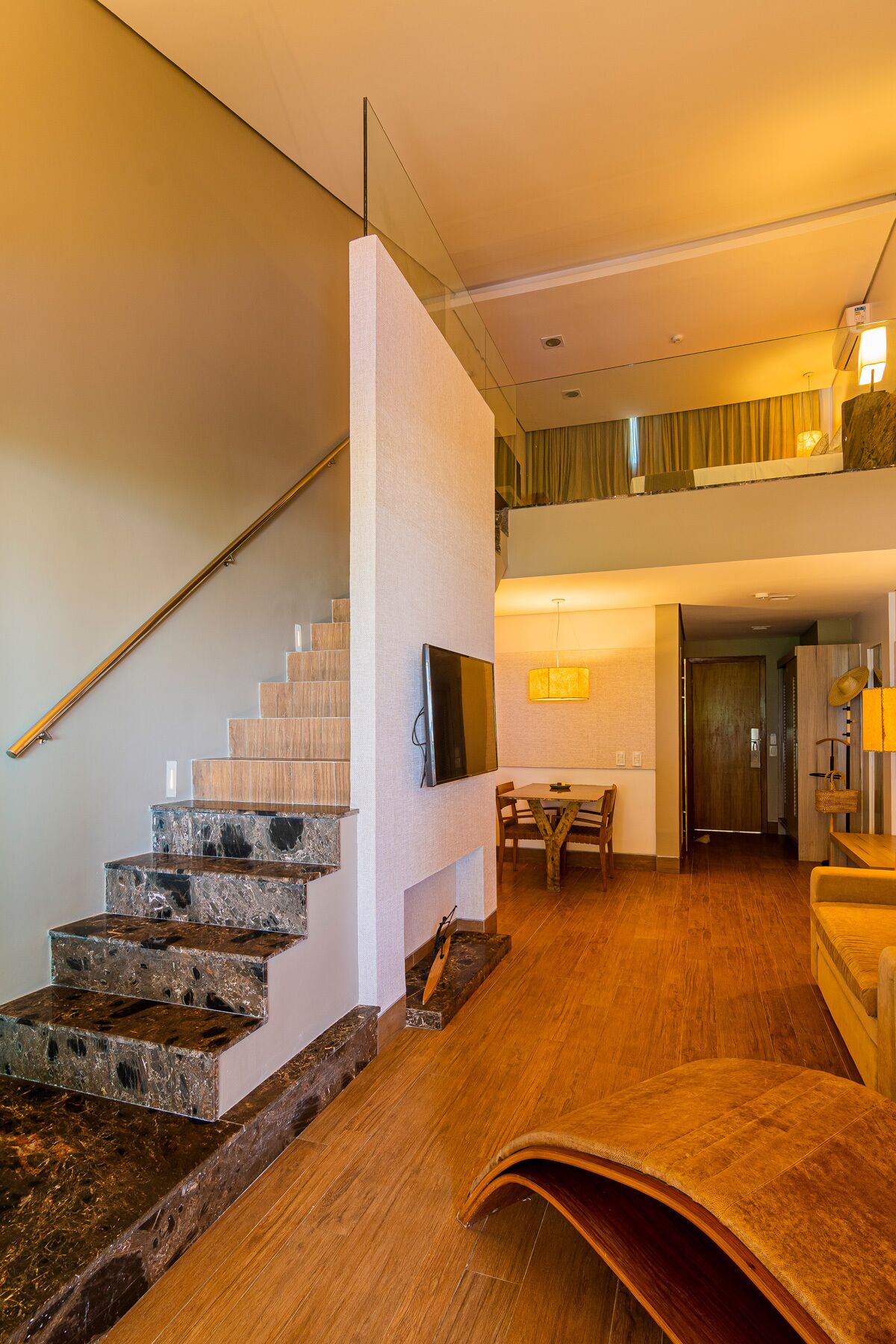 A modern interior with a wooden staircase, a flat-screen TV mounted on a wall, a dining area with a table and chairs, and a second-floor balcony with a view.