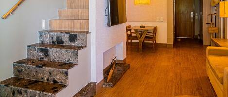 Un interior moderno con una escalera de madera, un televisor de pantalla plana montado en la pared, una zona de comedor con mesa y sillas, y un balcón en el segundo piso con vistas.