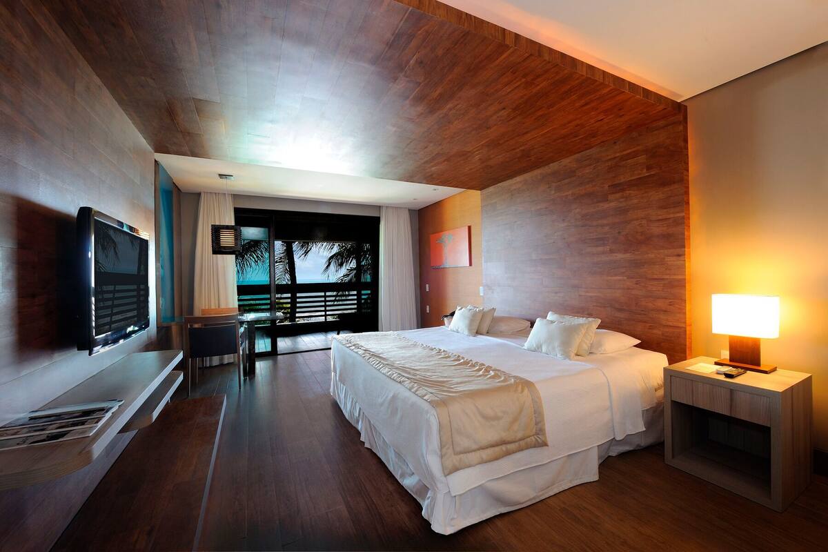Una habitación de hotel moderna con una cama grande, un televisor montado en la pared y un balcón con vistas a palmeras.