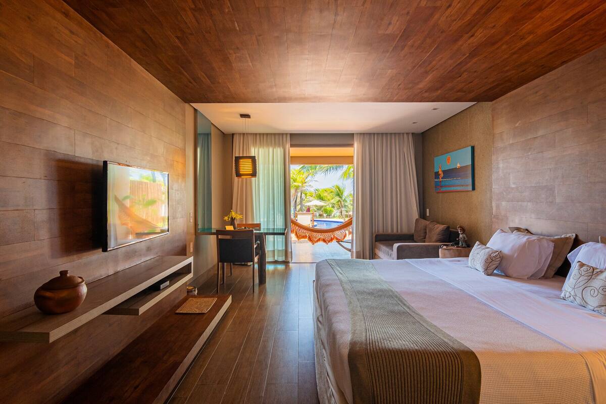 Habitación de hotel moderna con techo de madera, una cama grande, televisor, escritorio y vistas a una piscina y palmeras.