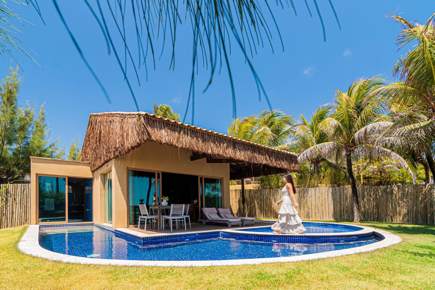 A modern house with a pool, a thatched roof, and palm trees.