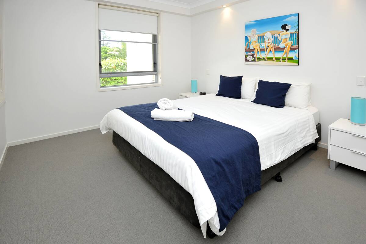 A neatly made bed with a blue and white bedspread, a window with a view of greenery, and a painting on the wall.