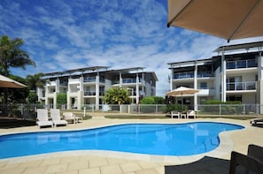 A swimming pool with lounge chairs, surrounded by a fenced area with a multi-story building in the background.