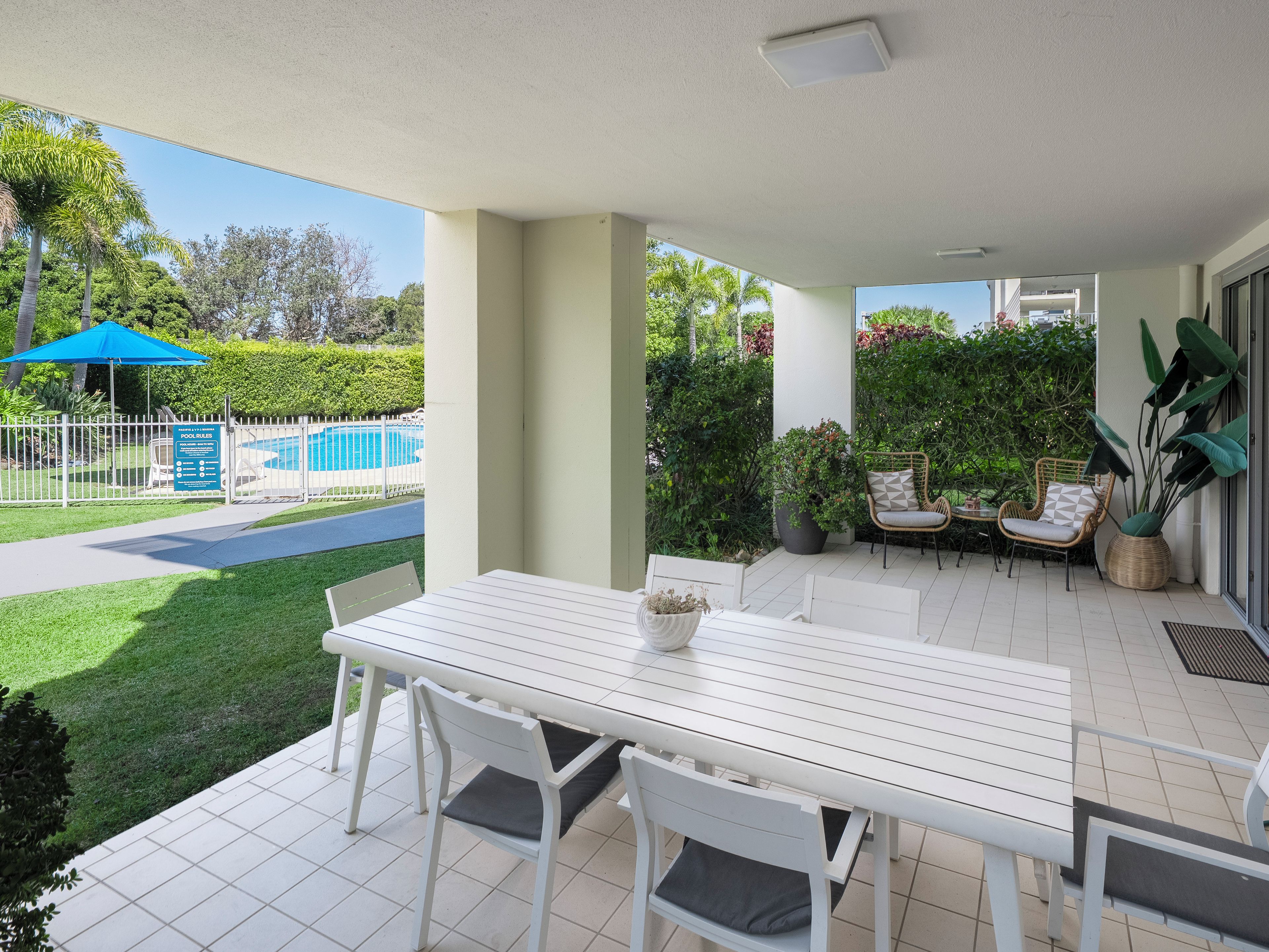 A covered patio area with a white table and chairs, a pool in the background, and a fenced garden.