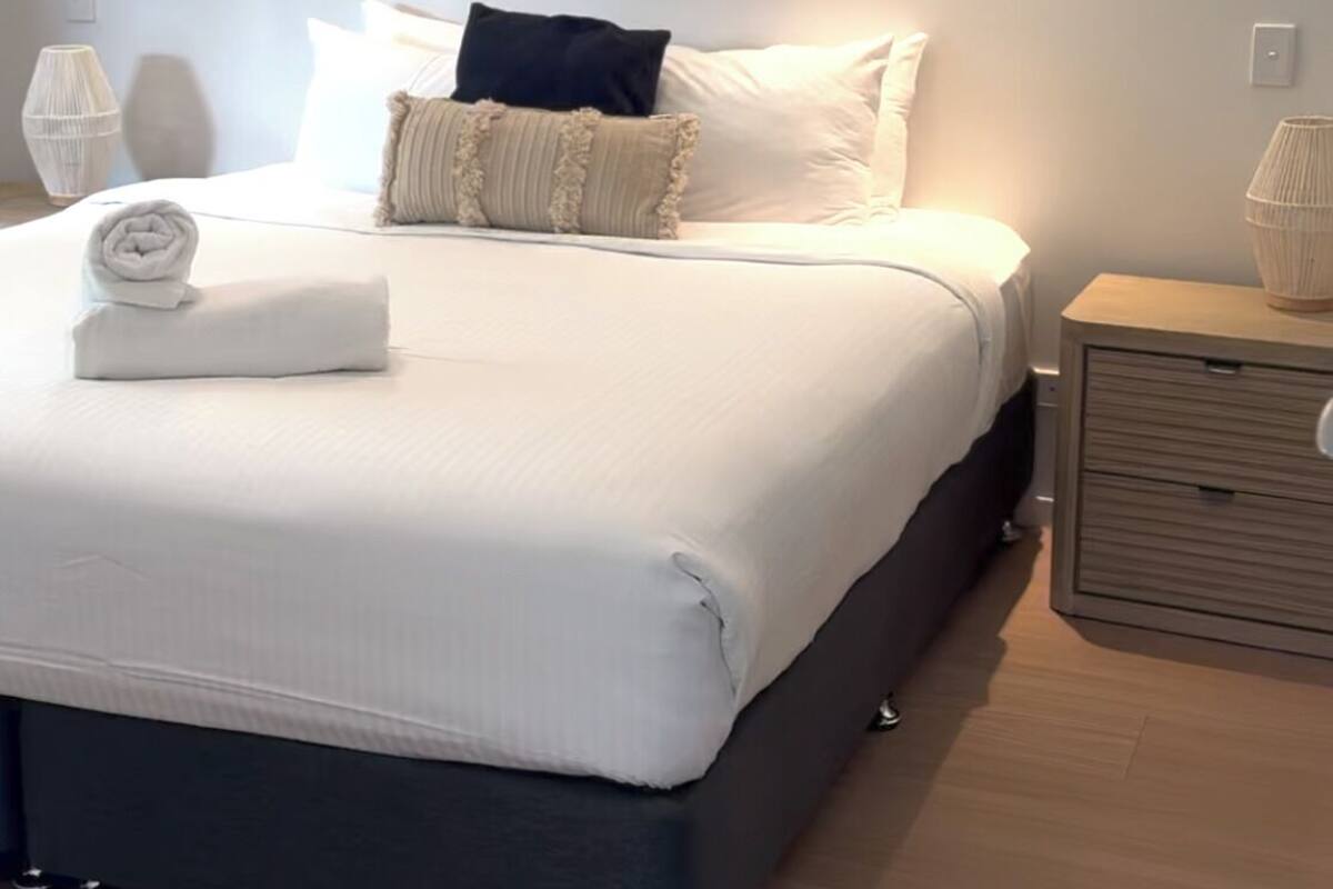 A neatly made bed with a rolled towel, a painting above it, and a bedside table with a lamp.