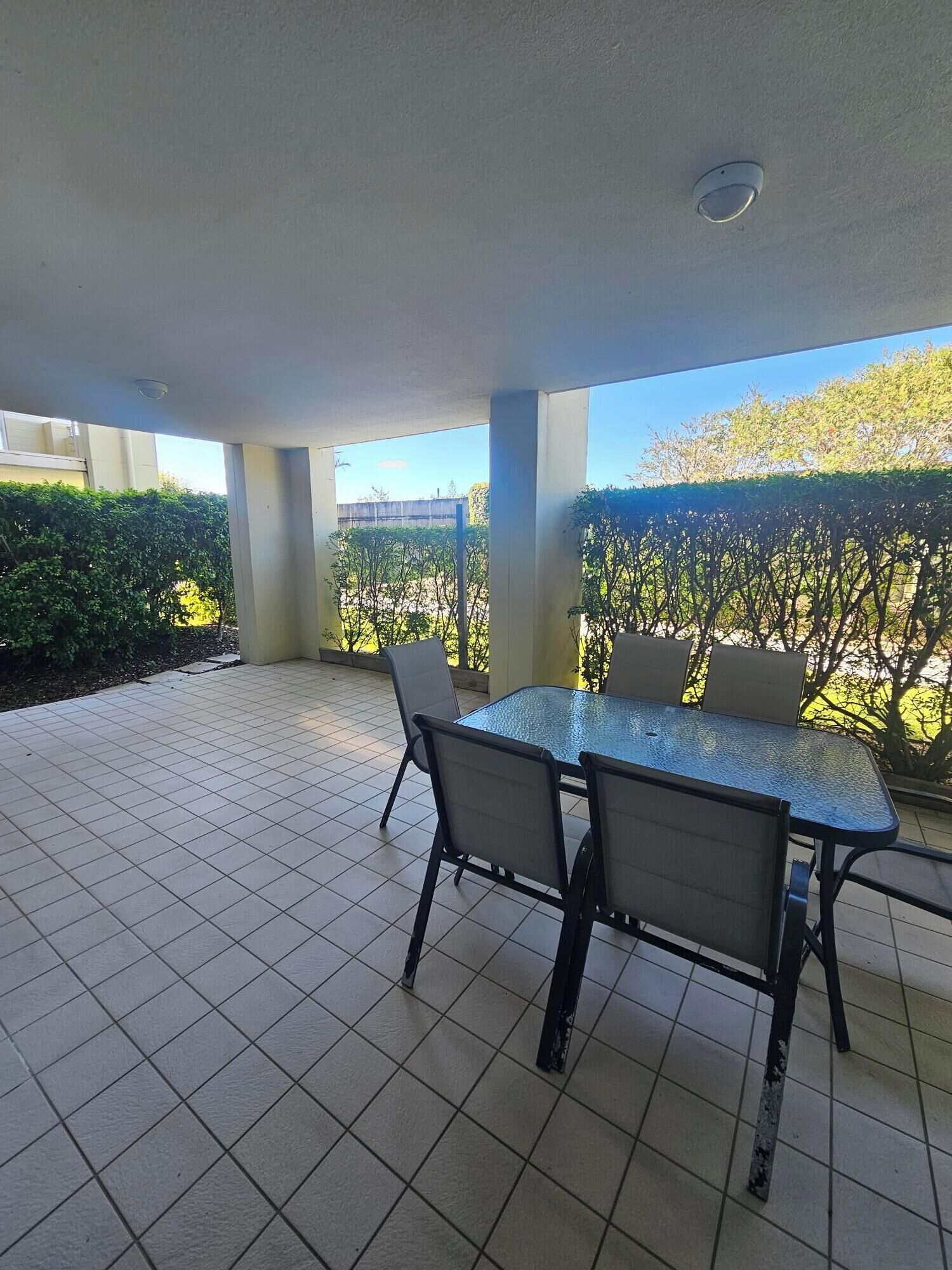Un espace extérieur aménagé en terrasse, doté d’un sol carrelé, d’une table à plateau en verre et de quatre chaises, et offrant une vue sur une haie et des arbres.