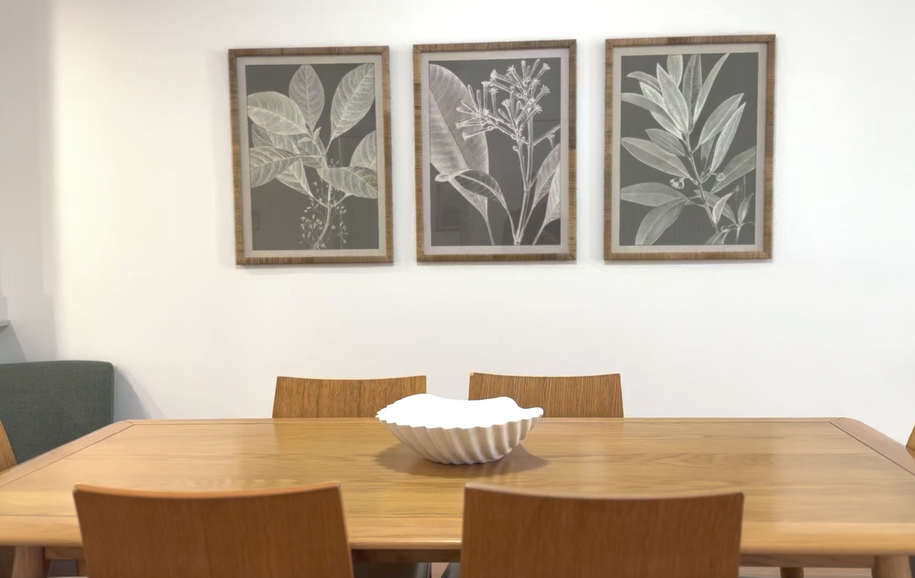 A dining table with a white bowl centerpiece, surrounded by six wooden chairs, with three framed botanical prints on the wall above.
