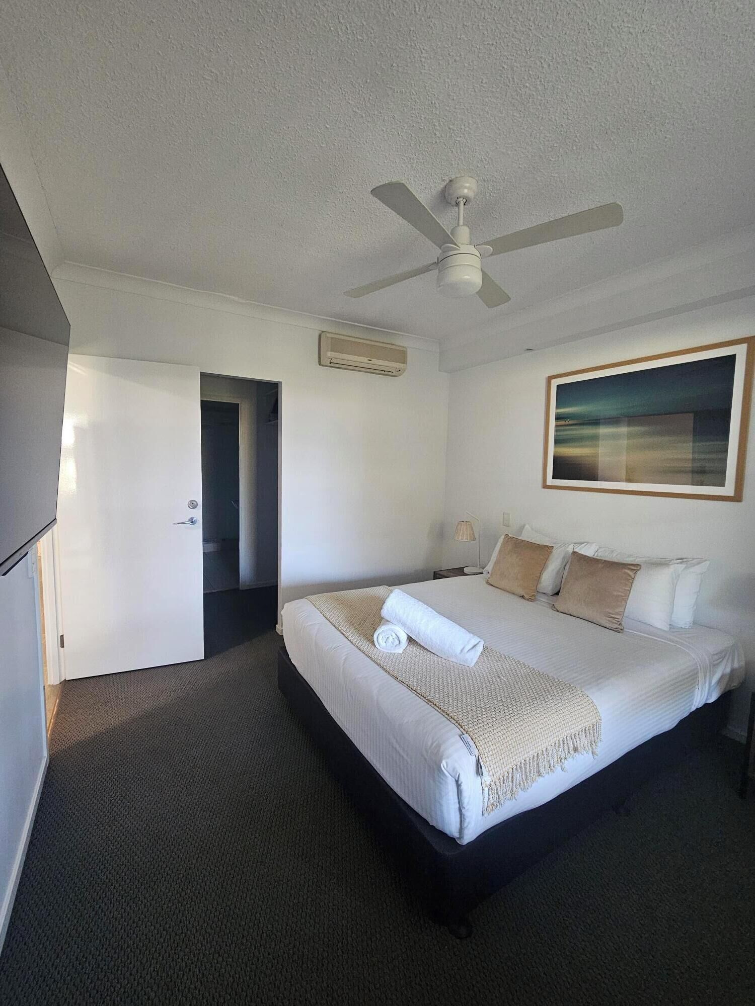 A hotel room with a bed, a ceiling fan, an air conditioning unit, a door, and a framed picture on the wall.