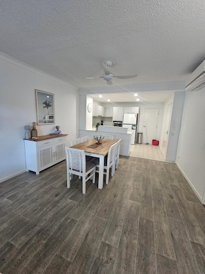 Una cocina moderna con armarios blancos, una zona de comedor con mesa de madera y sillas blancas, y un ventilador de techo.