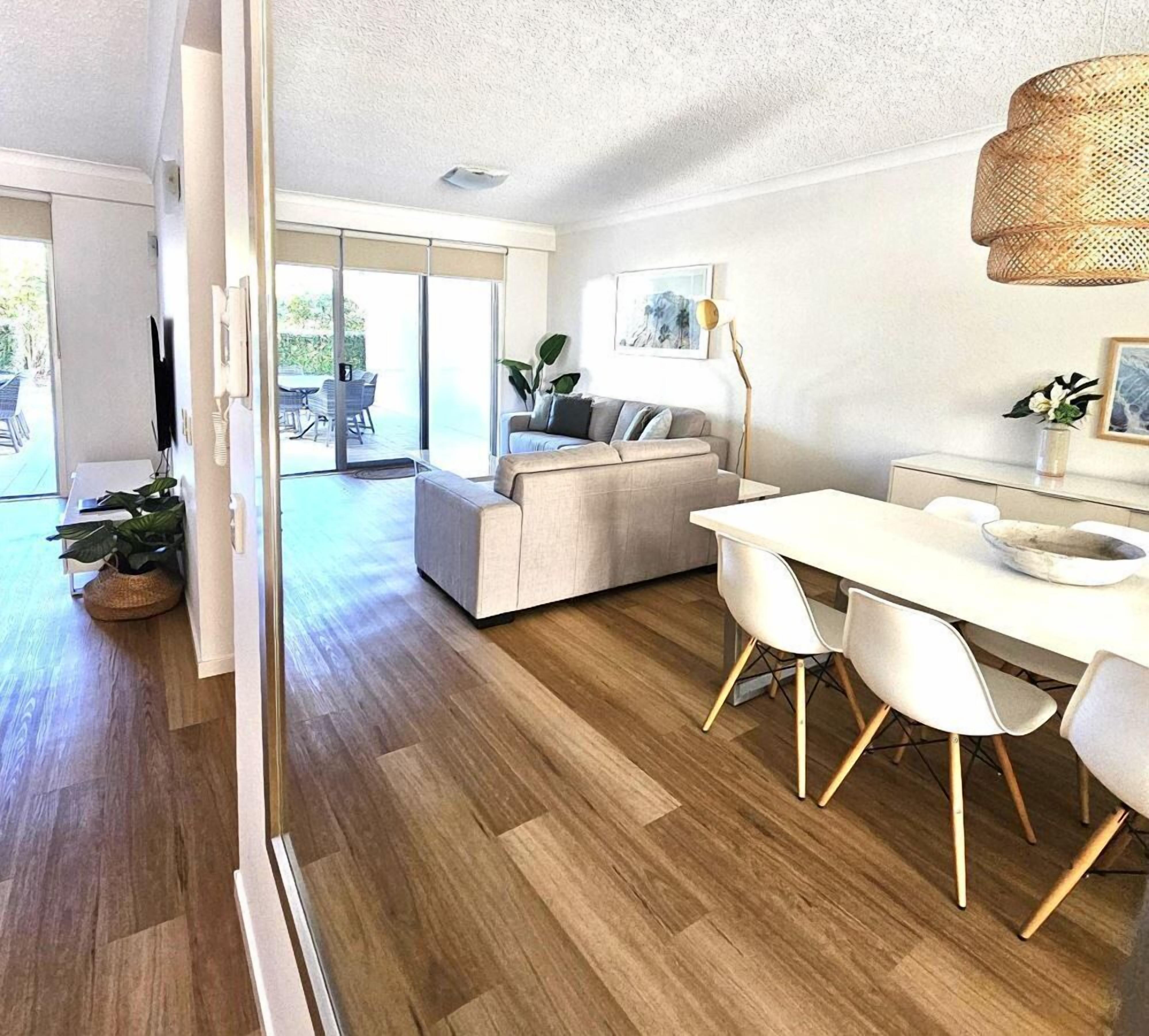 A modern living room with a sofa, dining table, and chairs. There is a large window providing natural light and a view of the outdoors.