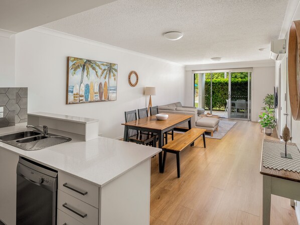 A modern kitchen with a dishwasher, a dining area with a wooden table and chairs, a sofa, and a large window with a view of greenery.