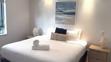 Una cama bien hecha con una toalla enrollada, un cuadro de una playa y una lámpara en una mesita de noche.