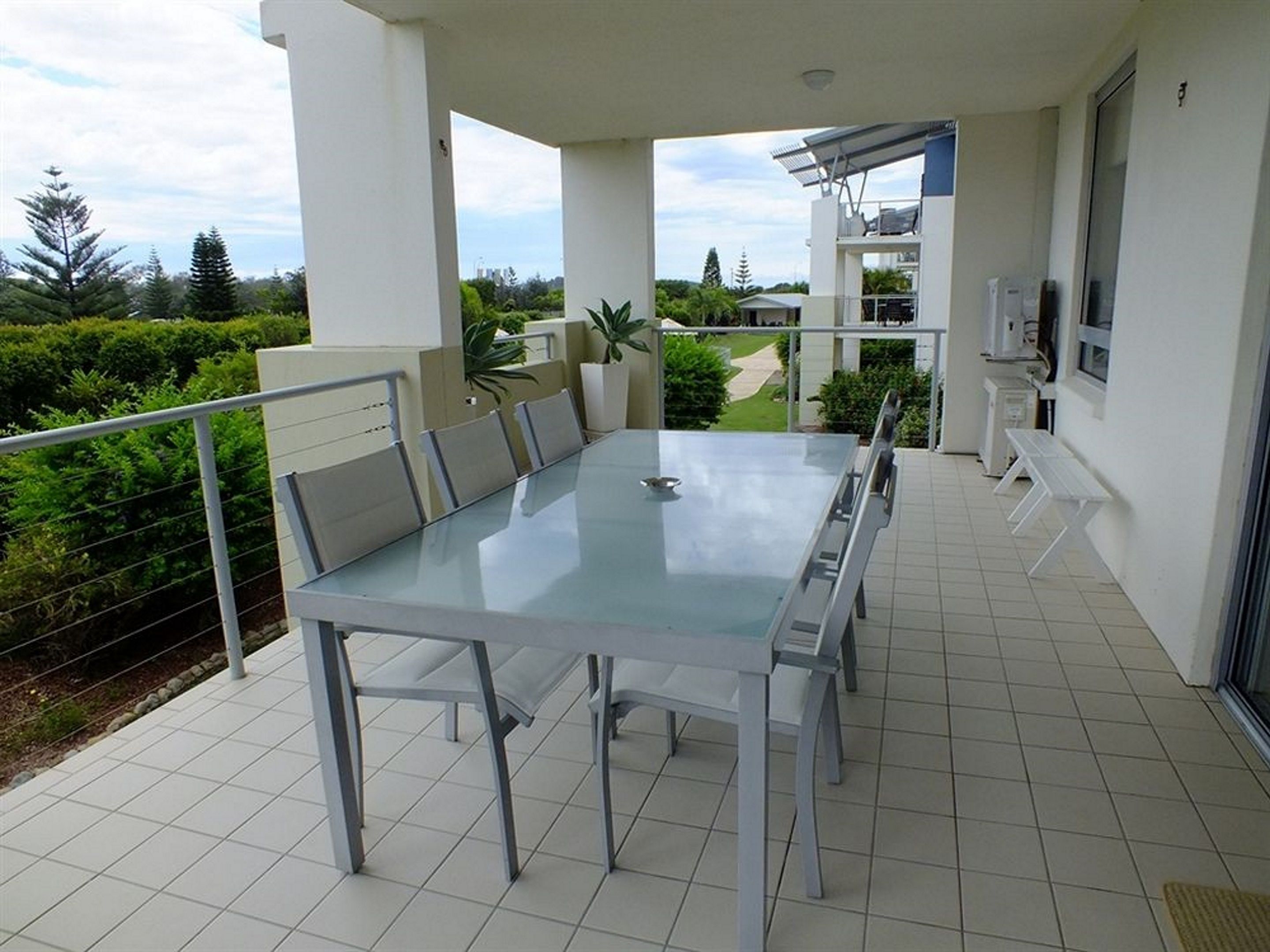 Un espace repas extérieur moderne, doté d’une table en verre et de chaises en métal, entouré d’une rambarde blanche et de plantes en pot.