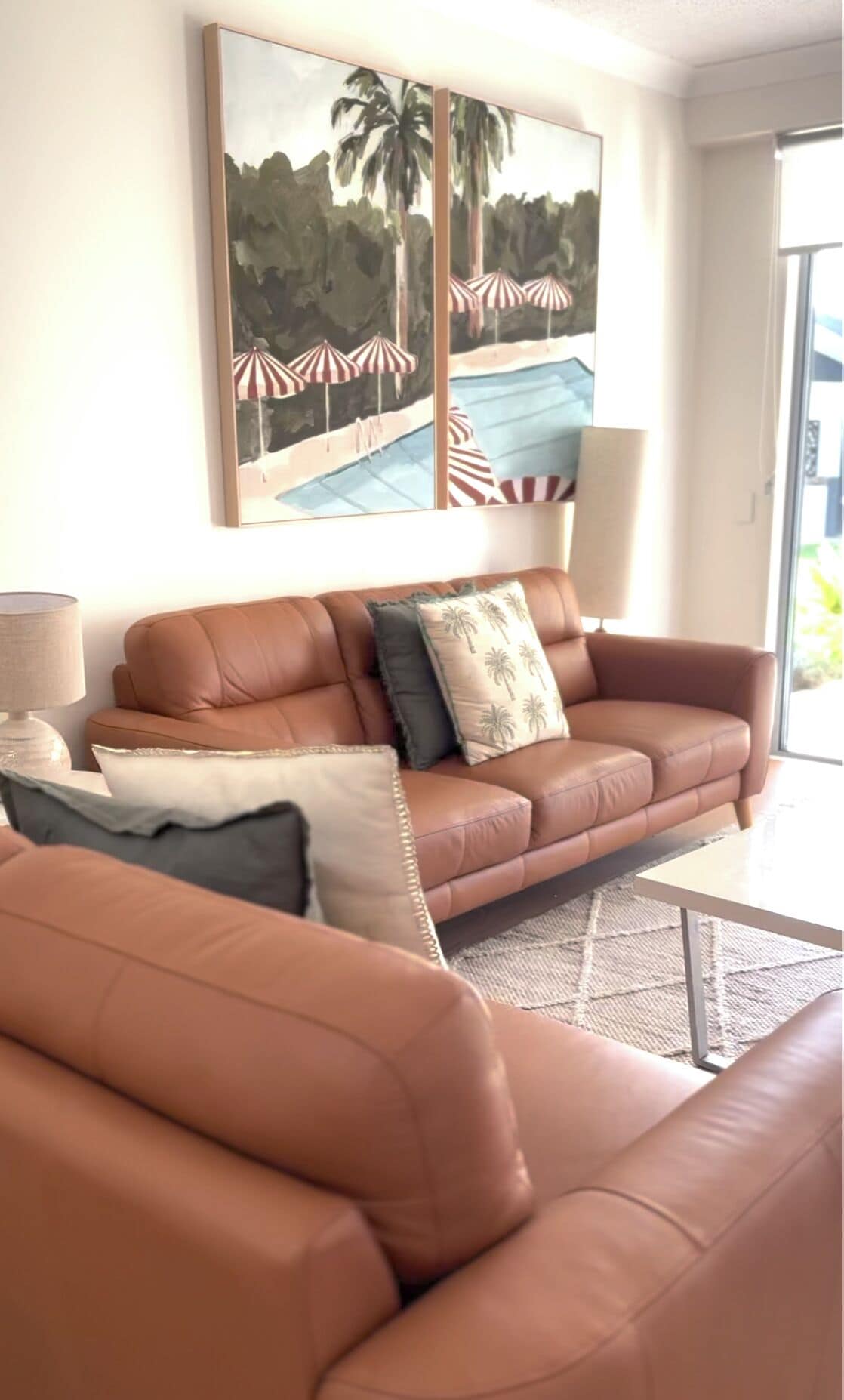 A living room with a brown leather sofa, a coffee table, and a painting of a beach scene.